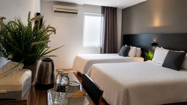 Habitación de hotel con dos camas, una mesita de noche con hervidor de agua, una bandeja con botellas de agua y una planta en maceta.