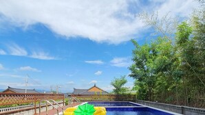 Una piscina con una valla de madera, un flotador inflable verde y edificios tradicionales al fondo.
