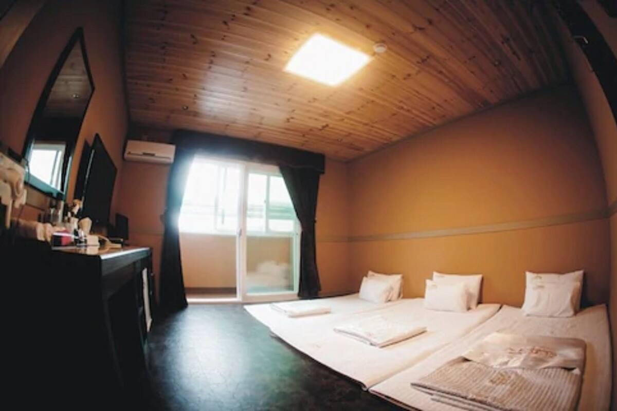 A room with a wooden ceiling, a bed with white bedding, a desk with a mirror, and a window with curtains.