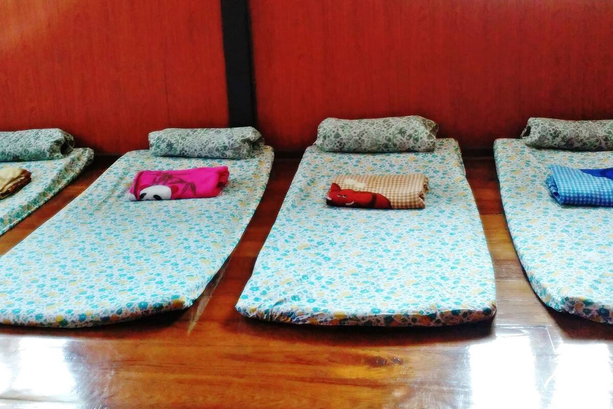 A row of single beds with floral-patterned bedding, a wooden floor, and a window with blinds.