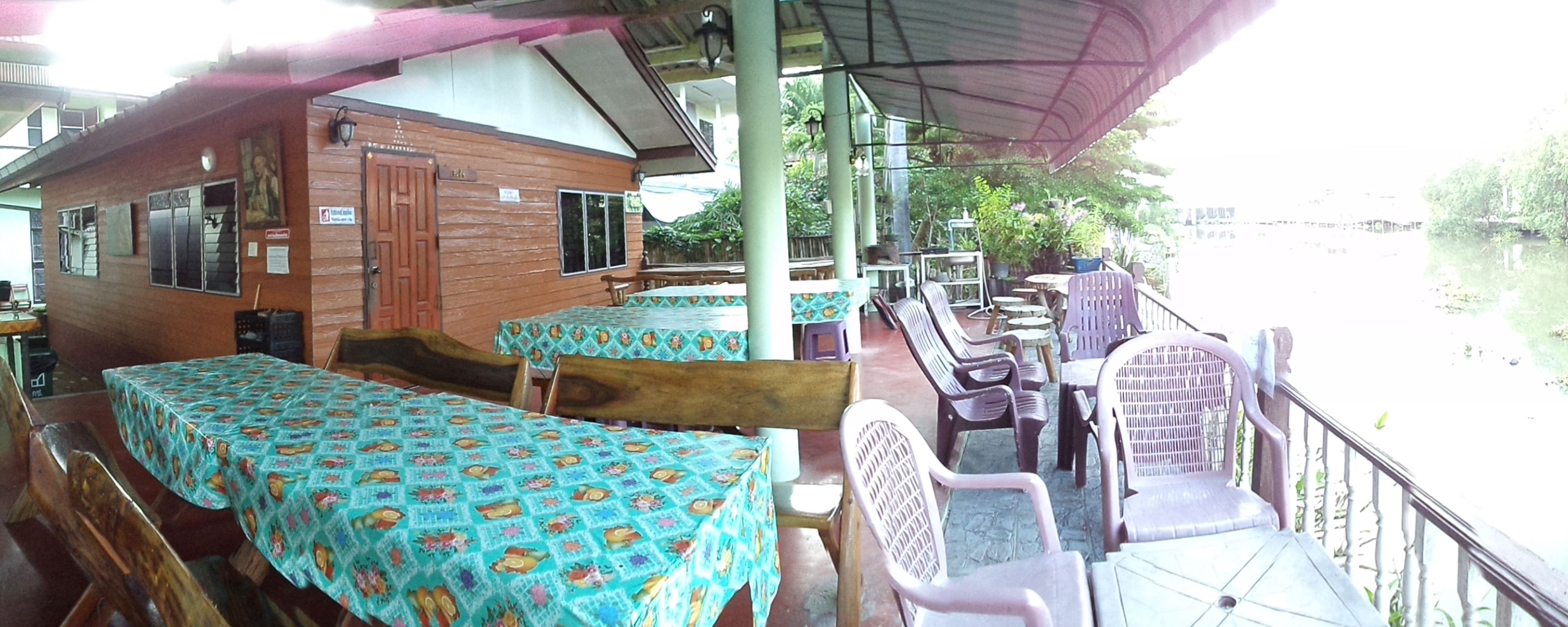 A riverside cabin with wooden walls and a covered patio area. There are several tables and chairs, some with colorful tablecloths.