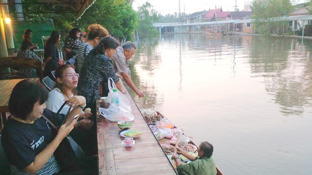 A long wooden boat with passengers and a table loaded with food items. - NR Homestay (Amphawa)