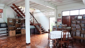 A spacious room with a wooden floor, a dining table with chairs, a staircase, a bookshelf, and a television. - NR Homestay (Amphawa)