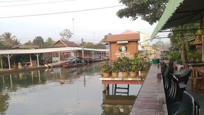 A riverside walkway with a small structure displaying a clock and contact information. - NR Homestay (Amphawa)