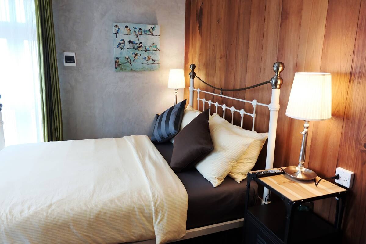 A neatly made bed with a white comforter and multiple pillows, a bedside table with a lamp, and a wall-mounted artwork.