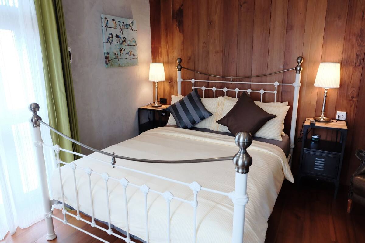 A neatly made bed with a white metal frame, two bedside tables with lamps, a wooden wall panel, and a painting on the wall.