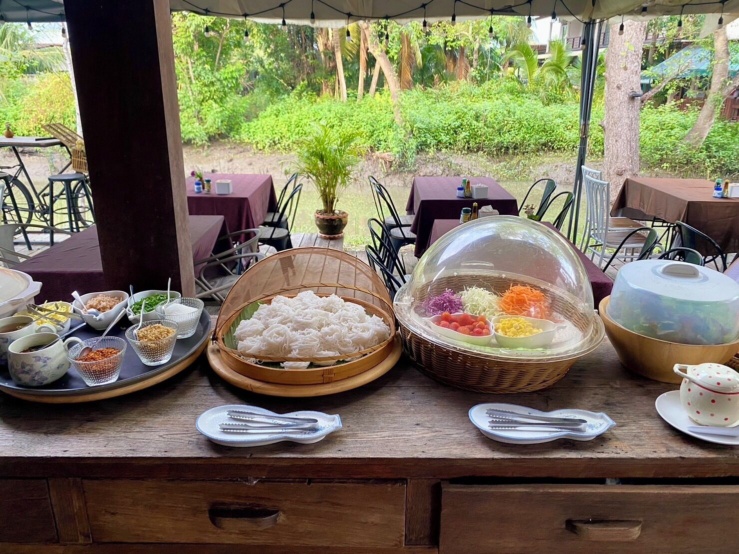 A buffet table with various dishes and utensils, set against a backdrop of lush greenery.