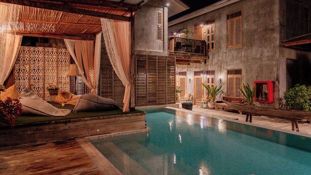 Un área de piscina con terraza de madera, un espacio de estar techado con cojines y un edificio con varias ventanas y balcones.