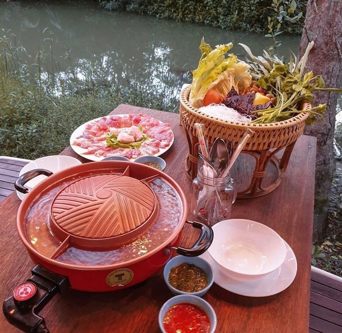 Uma preparação de panela quente com um caldeirão vermelho, uma cesta de legumes e um prato de carne fatiada sobre uma mesa de madeira perto da água.