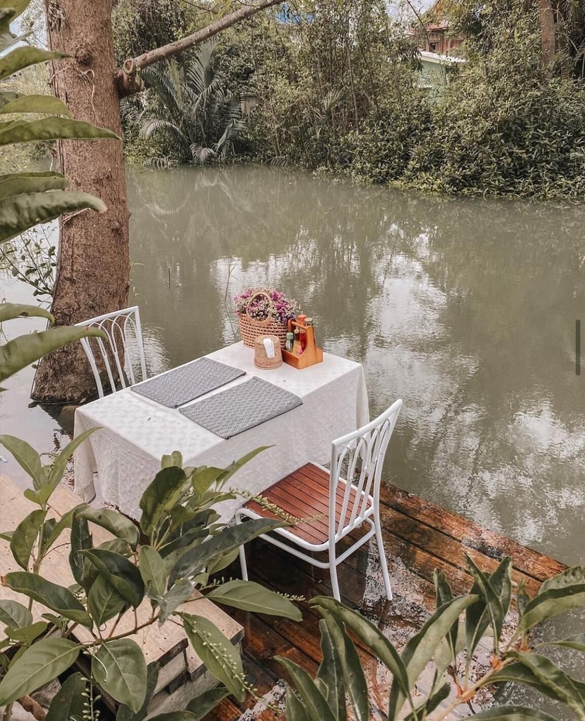 Una mesa junto a un río, con sillas, una cesta de flores y una pequeña construcción de madera.