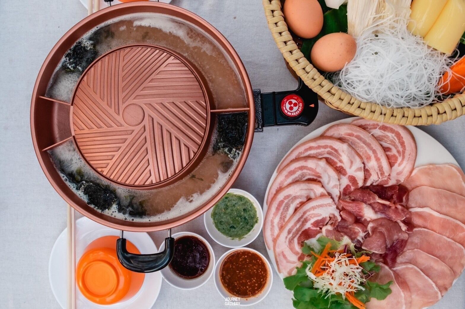 A hot pot setup with a ceramic pot, a woven basket with eggs, a plate of sliced meat, and bowls of condiments.