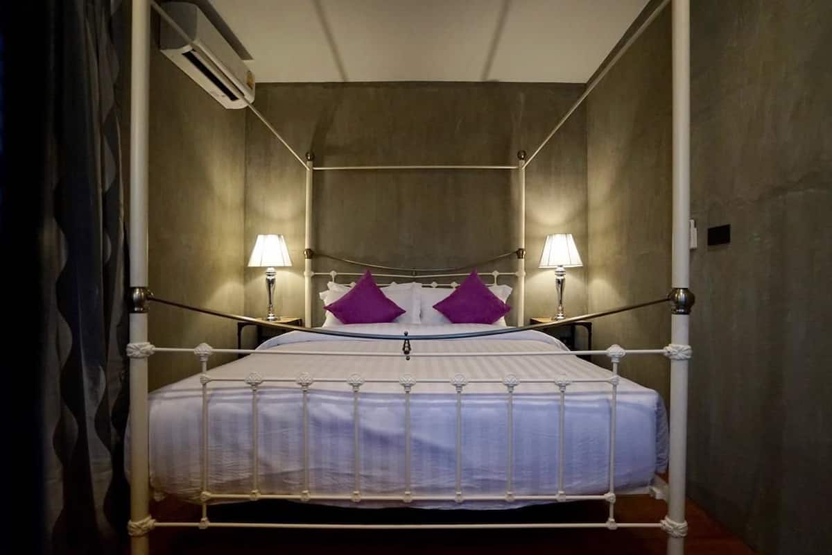 A four-poster bed with white metal frame and a white bedspread, two purple pillows, and two bedside lamps.