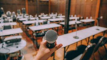 Una mano sostiene un micrófono en una sala con varias mesas y sillas, lo que sugiere un espacio para conferencias o reuniones.