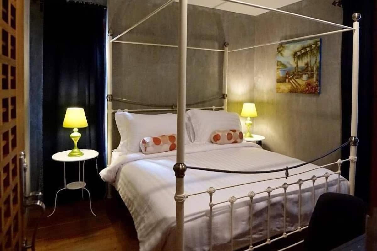 A four-poster bed with white bedding and polka dot pillows, a small bedside table with a lamp, and a painting on the wall.