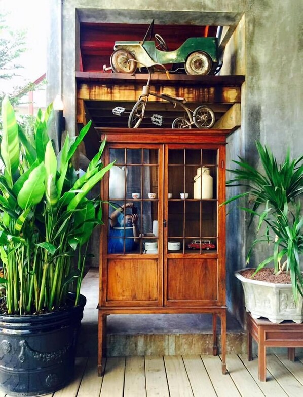 A wooden cabinet with glass doors, a green toy car on top, and potted plants on either side. - House of Passion Amphawa (Amphawa)