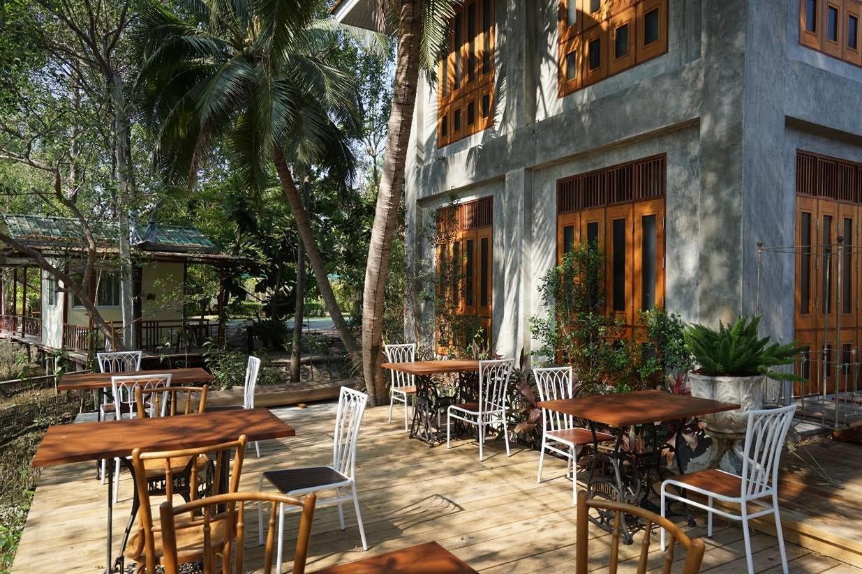 An outdoor patio with wooden tables and white chairs, surrounded by lush greenery and palm trees.