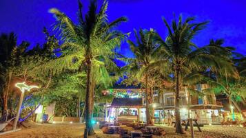 Un resort frente al mar con palmeras, mobiliario exterior y un edificio con señalización iluminada.