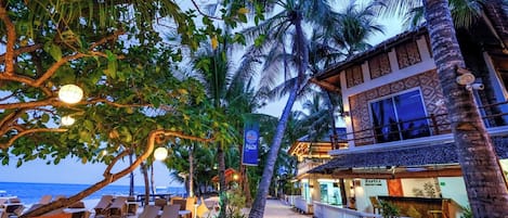 Un hotel frente a la playa con palmeras, sillas de descanso y un bar.