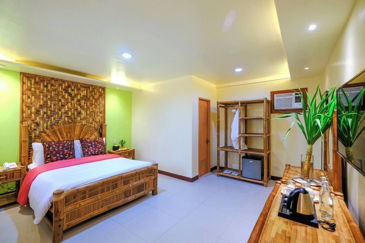 Une chambre avec un lit en bois, un téléviseur fixé au mur, une armoire en bois et une table de chevet avec une bouilloire et des verres.