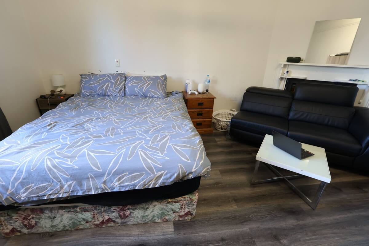 Um quarto com uma cama, uma mesa de cabeceira, um aparador, um sofá de couro preto e uma pequena mesa branca com um laptop.
