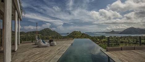 A wooden deck with a pool, outdoor seating, and a view of the ocean and mountains.