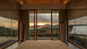 Une chambre avec un grand lit, une vue sur le coucher de soleil sur les montagnes et un balcon avec une balustrade.
