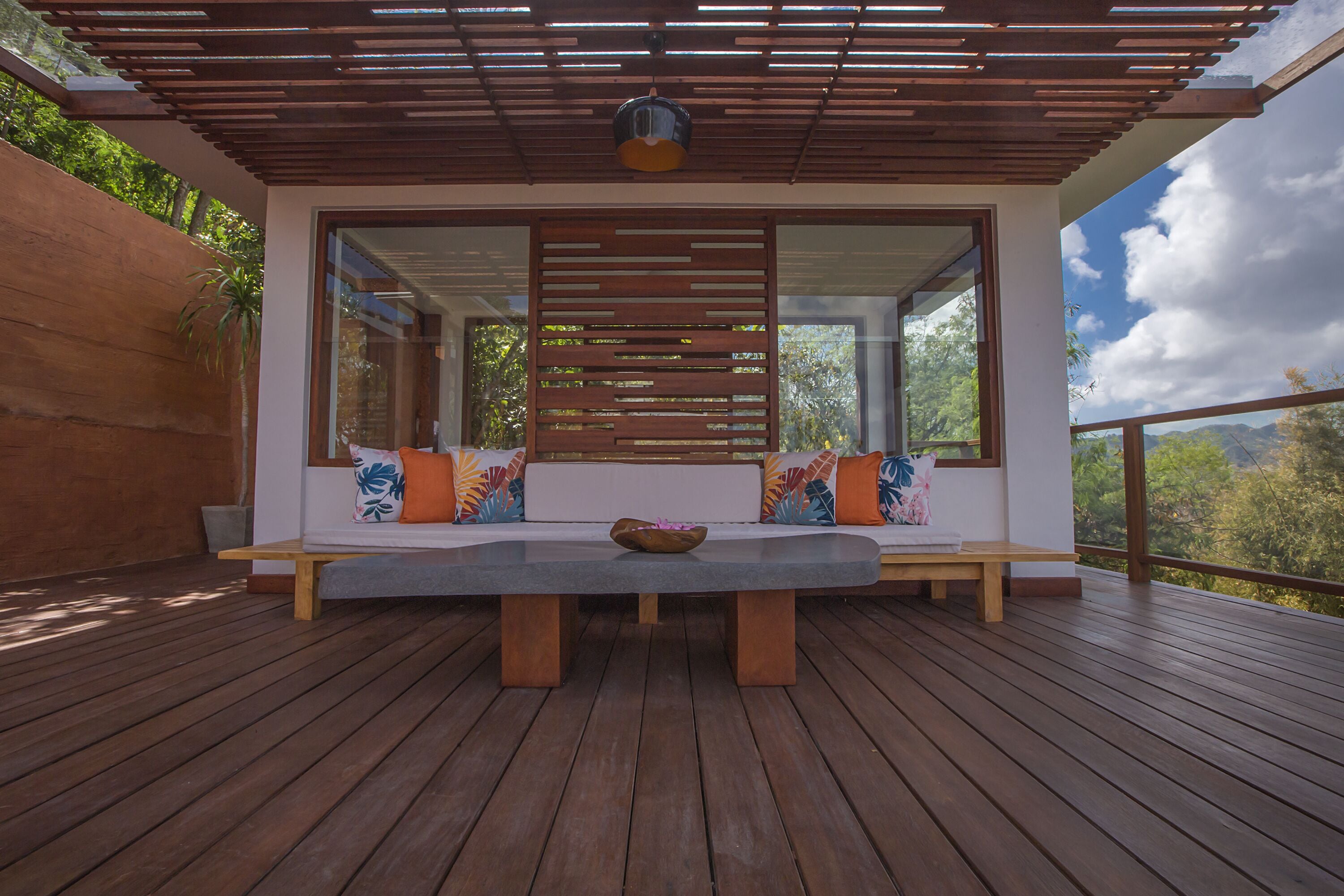 A wooden deck with a bench and cushions, a ceiling light, and a view of greenery.
