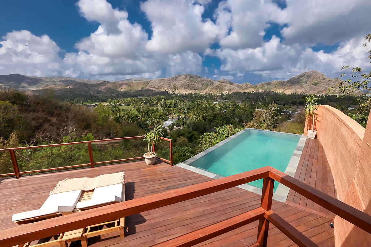 A wooden deck with a pool, lounge chairs, and a view of mountains and greenery.