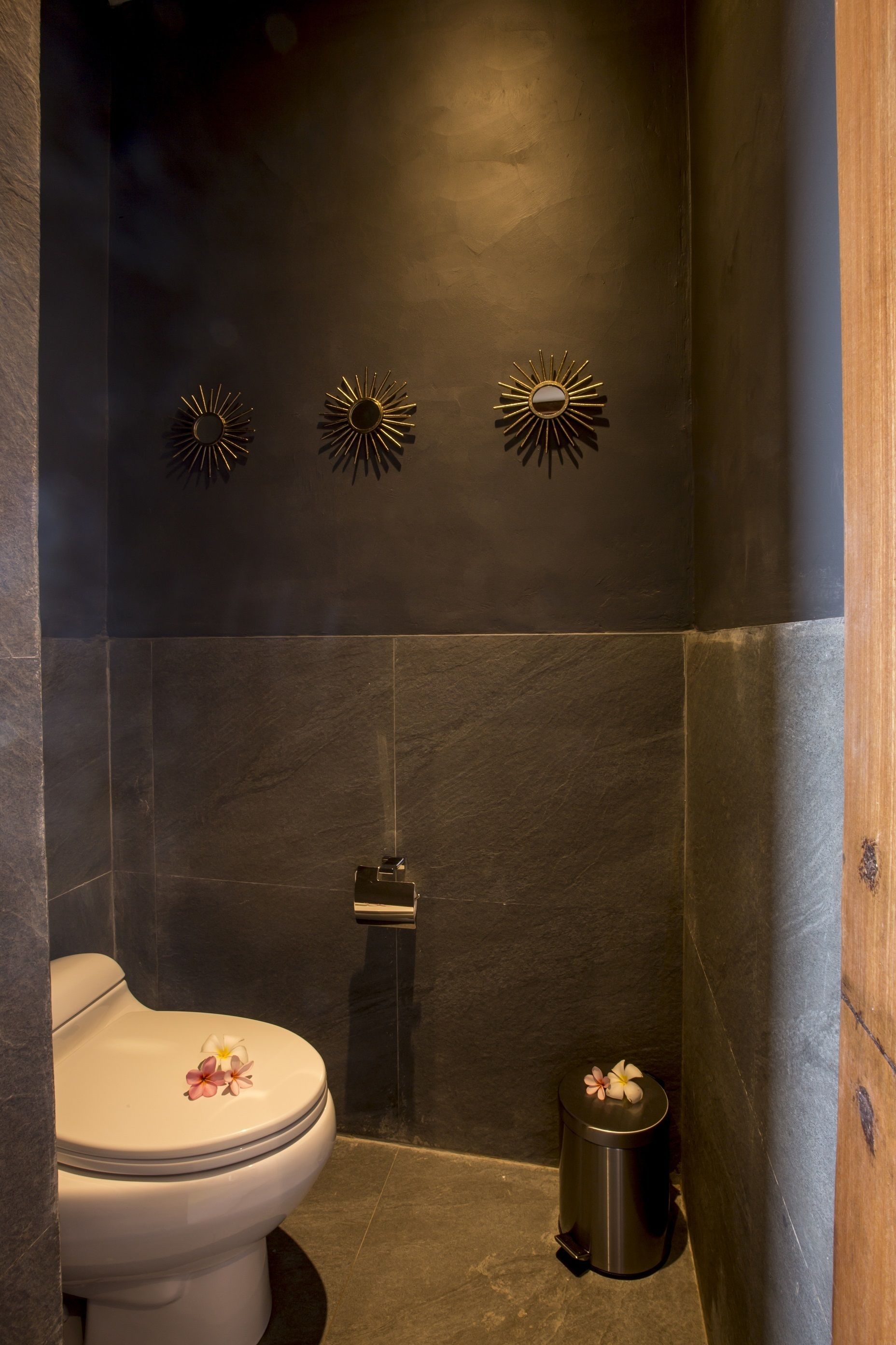 A modern bathroom with a dark tiled wall, a white toilet, and a trash can.