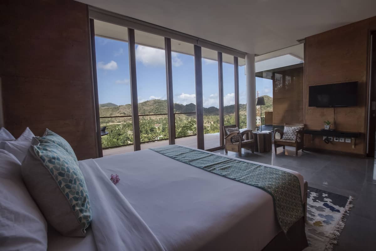 A modern hotel room with a large bed, a balcony with a view of the hills, a TV mounted on the wall, and a small seating area.