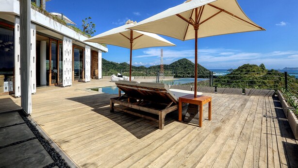 Une terrasse en bois avec deux parasols beiges, une chaise longue en bois et une piscine en arrière-plan.