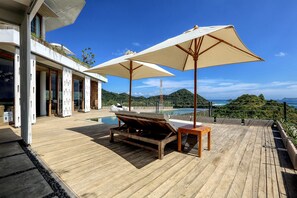 Una terraza de madera con dos sombrillas beige, una silla lounge de madera y una piscina al fondo.
