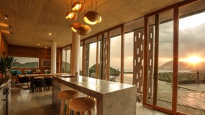 Une cuisine moderne dotée d’un grand îlot central, de tabourets en bois et offrant une vue sur le coucher de soleil derrière les montagnes.