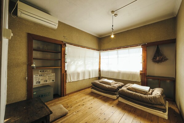 A room with a wooden floor, a bed with a white blanket, a small table, a shelf with a plant, and a sign about an emergency ladder.