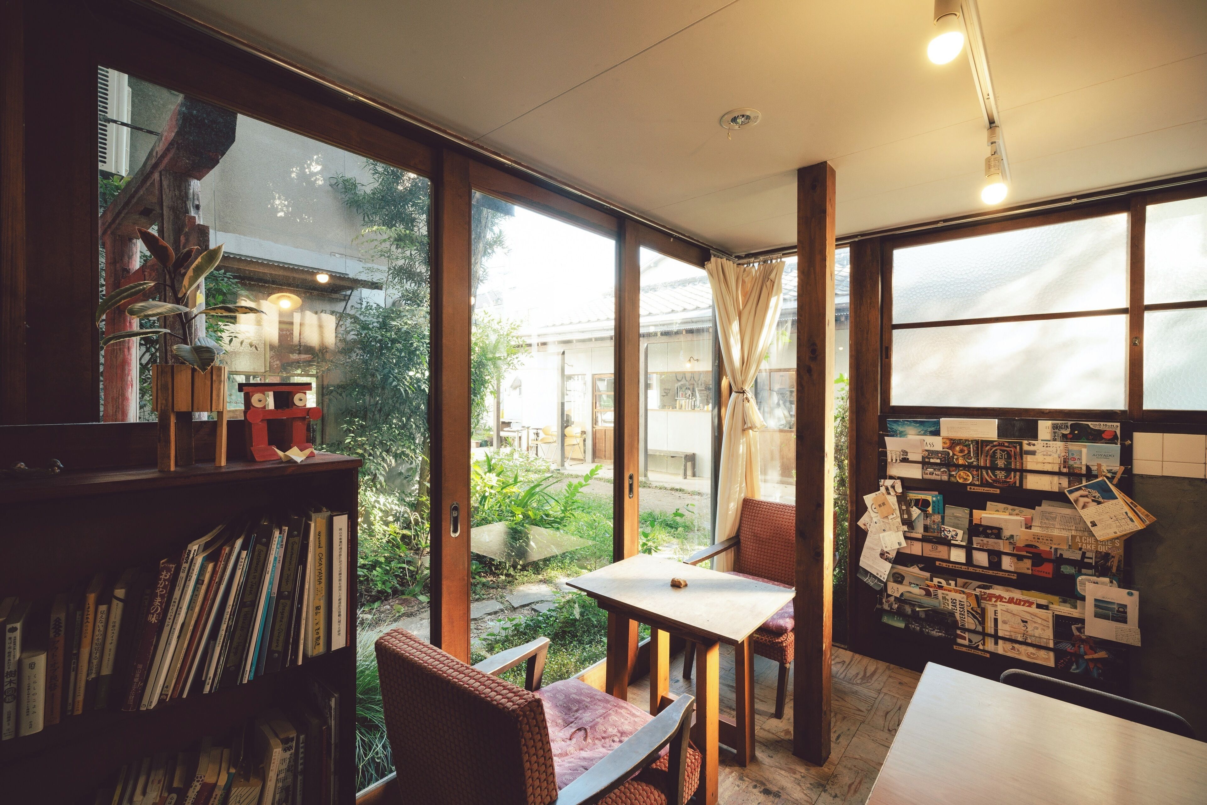 A room with a large window, a bookshelf, a table with a chair, and a bulletin board with papers.