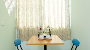 A room with a wooden table set for two, two blue chairs, a floral curtain, and a wall-mounted light fixture. - The Garden Place (Nonthaburi)