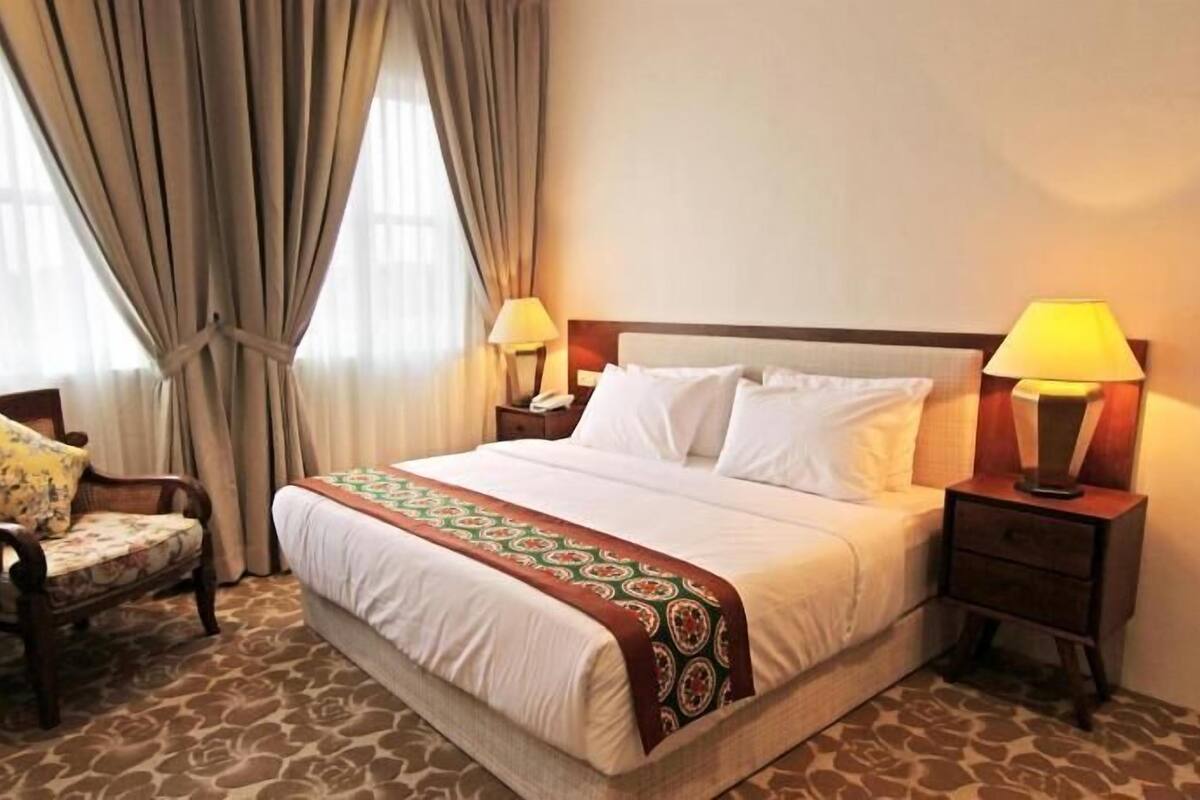 Una camera d'albergo con un letto, due abat-jour, una poltrona con cuscino e un tappeto decorato.
