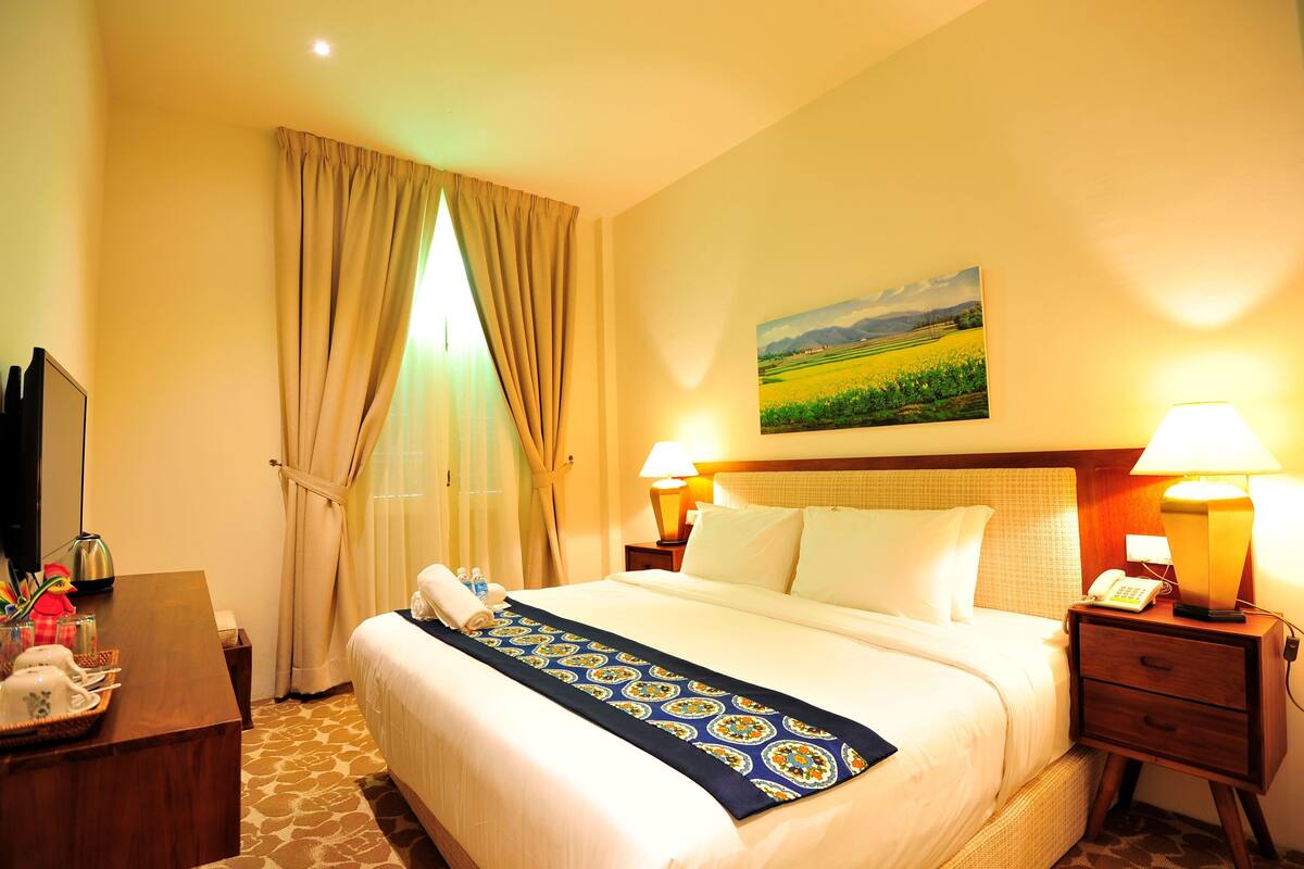 Una camera d'albergo con un letto, un comodino con una lampada, un quadro al muro e una finestra con tende.