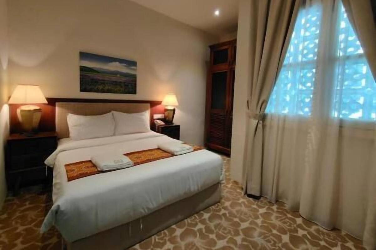 Camera d'albergo con un letto, due abat-jour, un quadro appeso al muro e una finestra con tende.