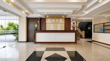 A hotel reception area with a reception desk, clocks, a staircase, and a glass door leading outside.