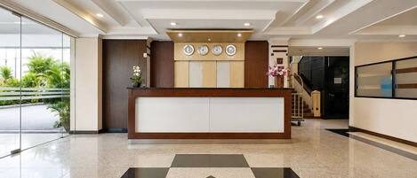 A hotel reception area with a reception desk, clocks, a staircase, and a glass door leading outside.