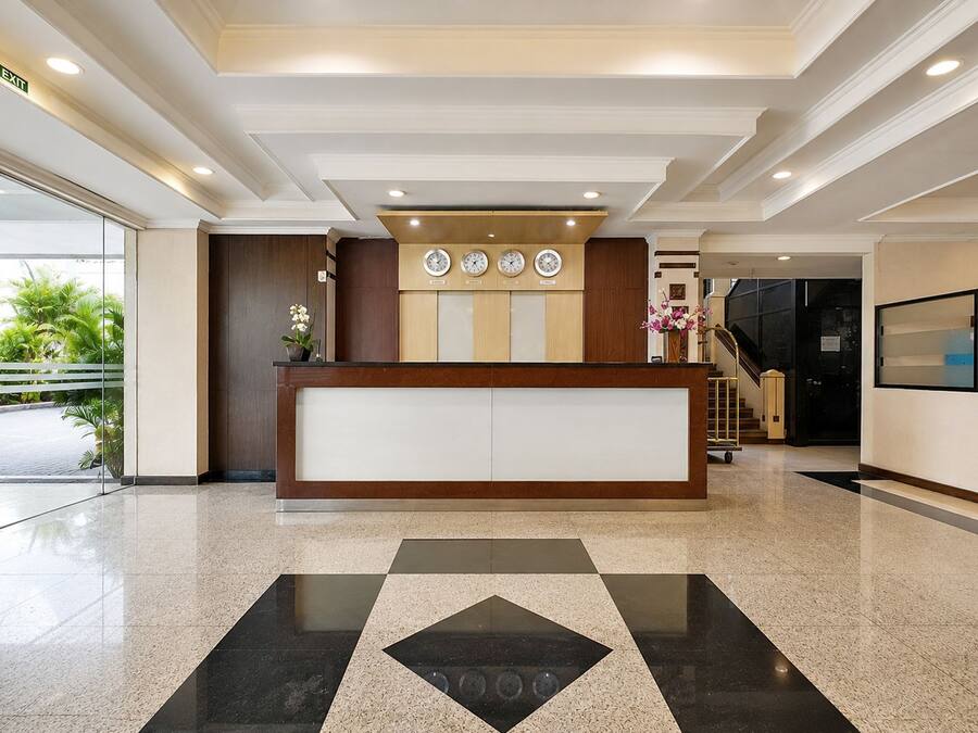 A hotel reception area with a reception desk, clocks, a staircase, and a glass door leading outside.