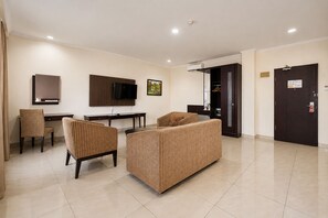 A hotel room with a flat-screen TV, a desk, two chairs, a sofa, and a door with a security lock. - Palette Citra Inn (Cikarang)