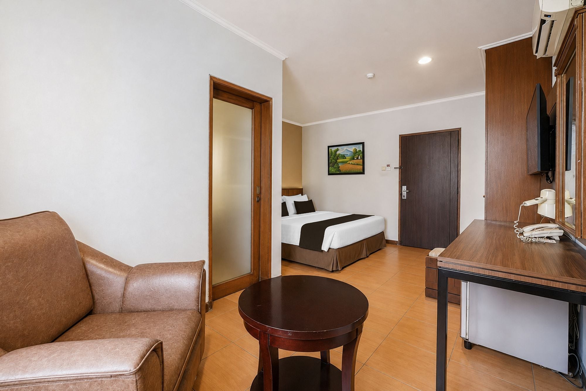 A hotel room with a sofa, a round table, a bed with a headboard, a painting on the wall, and a door leading to another room.