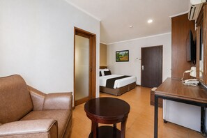 A hotel room with a sofa, a round table, a bed with a headboard, a painting on the wall, and a door leading to another room. - Palette Citra Inn (Cikarang)
