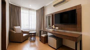 A hotel room with a sofa, a small table, a desk with a flat-screen TV, a mini-fridge, and an air conditioning unit.