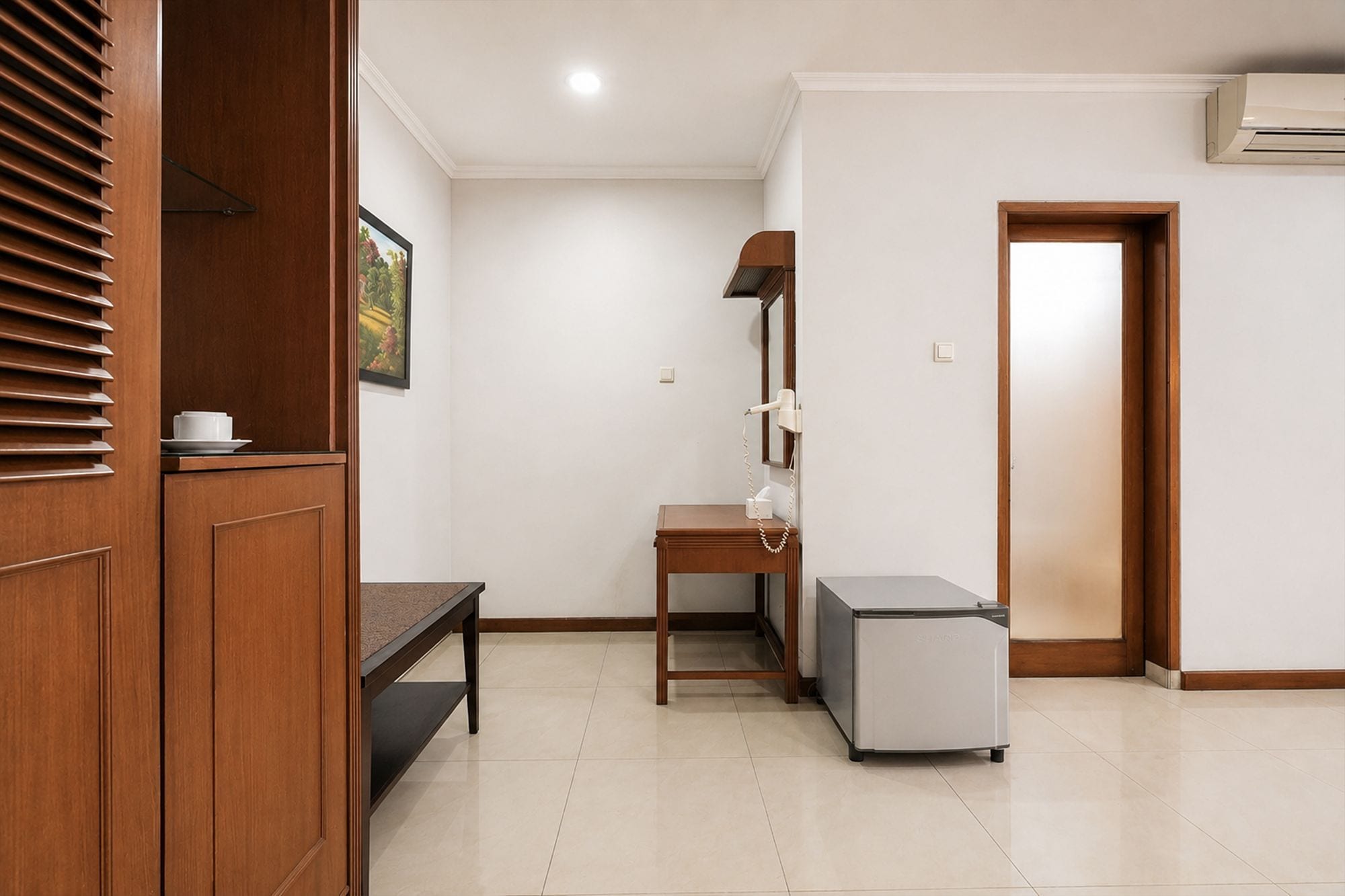 A hotel room with a wooden wardrobe, a desk with a chair, a small refrigerator, and a bedside table.