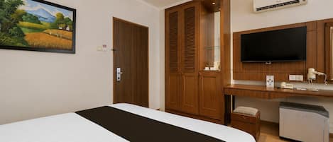 A hotel room with a bed, a desk with a flat-screen TV, a wardrobe, and an air conditioning unit.