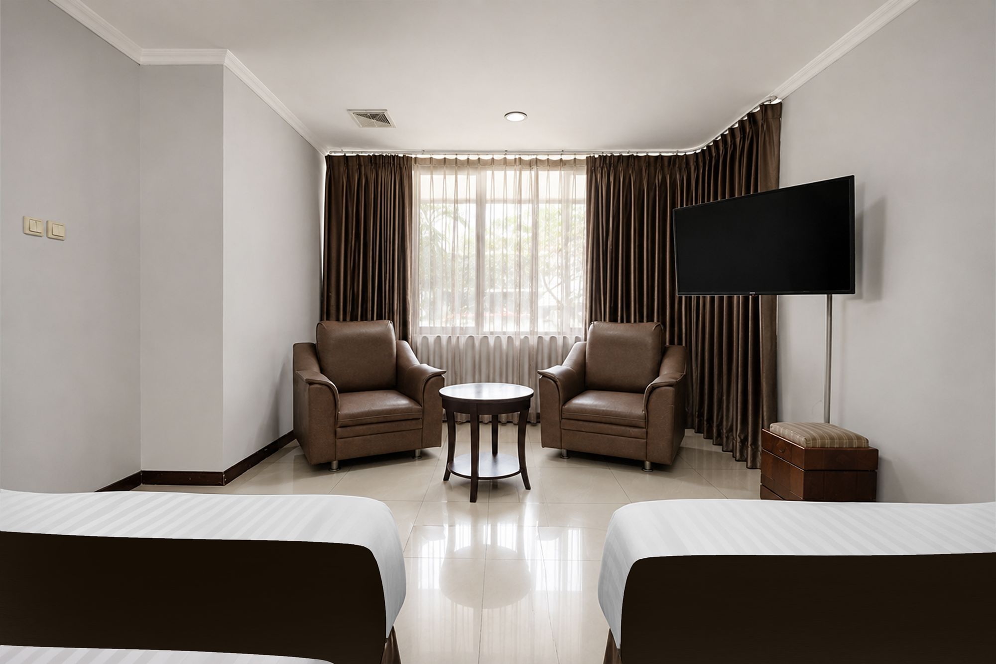 Una habitación de hotel con dos camas, televisión, dos sillones, una mesita y ventana con cortinas.