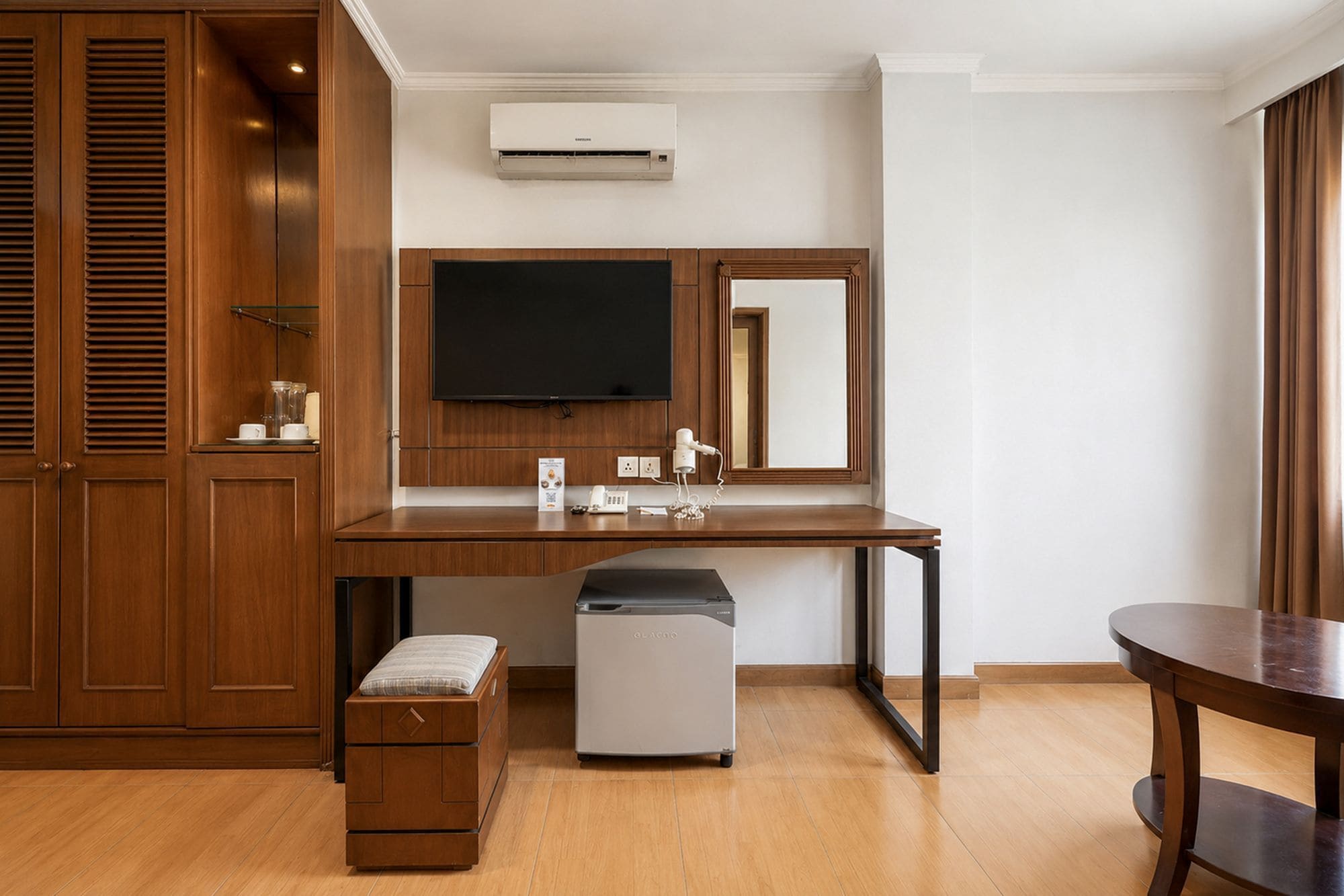 A hotel room with a wooden wardrobe, a desk with a hairdryer, a small fridge, a flat-screen TV, and a mirror.